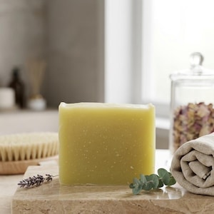 May include: A rectangular bar of pale green soap with visible small white flecks throughout its surface. The soap is positioned upright on a polished beige stone slab, accompanied by a small sprig of lavender and a single eucalyptus stem.