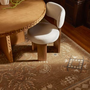 May include: A round light oak dining table with a decorative studded edge and a matching chair featuring a white upholstered seat and backrest, positioned on a brown area rug with a geometric pattern and a white vase with green foliage.