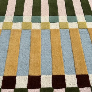 May include: A rectangular area rug featuring a geometric pattern of vertical stripes and squares in a palette of mustard yellow, light blue, olive green, soft pink, cream, and dark navy blue, with a textured, tufted pile construction throughout.
