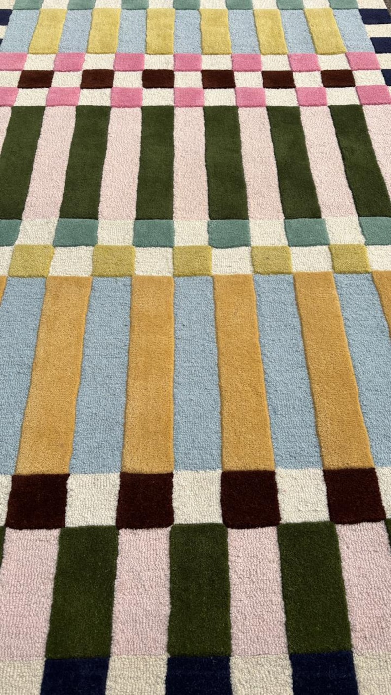 May include: A rectangular area rug featuring a geometric pattern of vertical stripes and squares in a palette of mustard yellow, light blue, olive green, soft pink, cream, and dark navy blue, with a textured, tufted pile construction throughout.