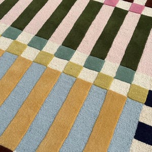 May include: A rectangular area rug featuring a geometric grid pattern of vertical and horizontal stripes. The design incorporates a palette of mustard yellow, light blue, olive green, soft pink, cream, and dark navy blue in a woven texture.