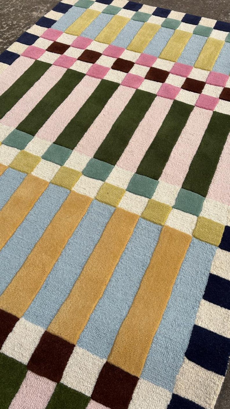 May include: A rectangular area rug featuring a geometric grid pattern of vertical and horizontal stripes. The design incorporates a palette of mustard yellow, light blue, olive green, soft pink, cream, and dark navy blue in a woven texture.