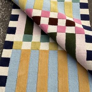 May include: A rectangular tufted rug featuring a geometric pattern of squares and vertical stripes in navy blue, mustard yellow, light blue, sage green, pink, dark brown, and cream. One corner of the rug is folded over to reveal the underside texture.