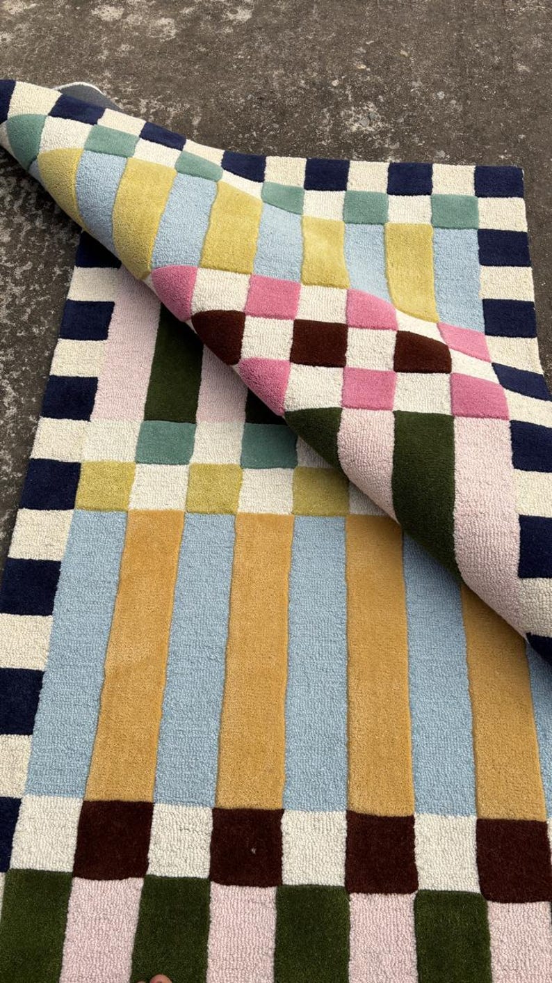 May include: A rectangular tufted rug featuring a geometric pattern of squares and vertical stripes in navy blue, mustard yellow, light blue, sage green, pink, dark brown, and cream. One corner of the rug is folded over to reveal the underside texture.