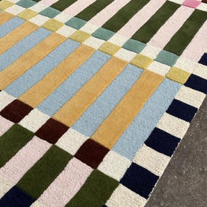 May include: A rectangular area rug featuring a grid pattern of thick, vertical and horizontal stripes in various colors including navy blue, olive green, light pink, mustard yellow, pale blue, and cream, with a textured, tufted surface finish.