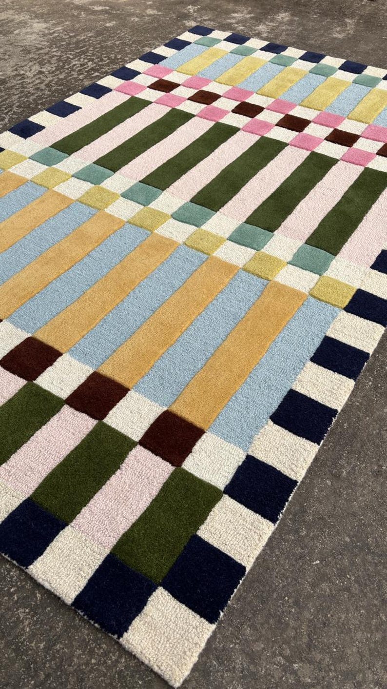 May include: A rectangular area rug featuring a grid pattern of thick, vertical and horizontal stripes in various colors including navy blue, olive green, light pink, mustard yellow, pale blue, and cream, with a textured, tufted surface finish.
