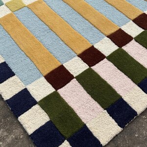 May include: A tufted area rug featuring a geometric pattern of rectangular blocks in shades of mustard yellow, light blue, olive green, dark navy, deep brown, soft pink, and cream. The textured surface displays a repeating grid-like arrangement.