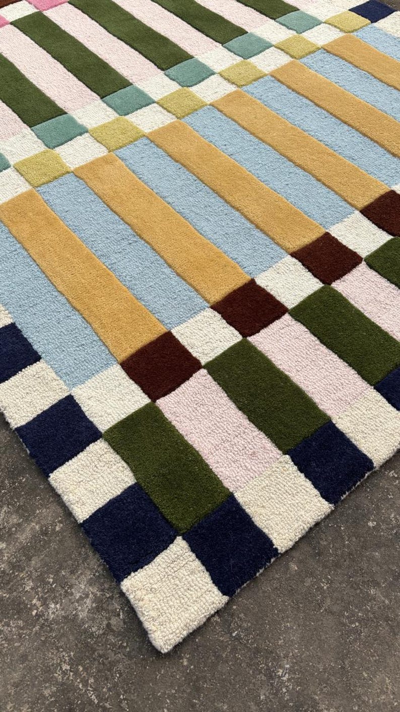 May include: A tufted area rug featuring a geometric pattern of rectangular blocks in shades of mustard yellow, light blue, olive green, dark navy, deep brown, soft pink, and cream. The textured surface displays a repeating grid-like arrangement.