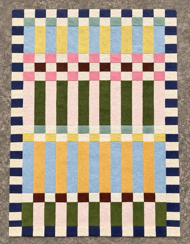 May include: A rectangular area rug featuring a geometric pattern of vertical stripes and horizontal bands. The design incorporates a palette of navy blue, cream, light blue, yellow, pink, dark brown, olive green, and soft teal in a grid layout.