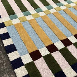May include: A rectangular area rug featuring a geometric pattern of alternating vertical stripes and squares. The design incorporates a palette of navy blue, olive green, mustard yellow, light blue, pale pink, cream, and lime green tufted textures.