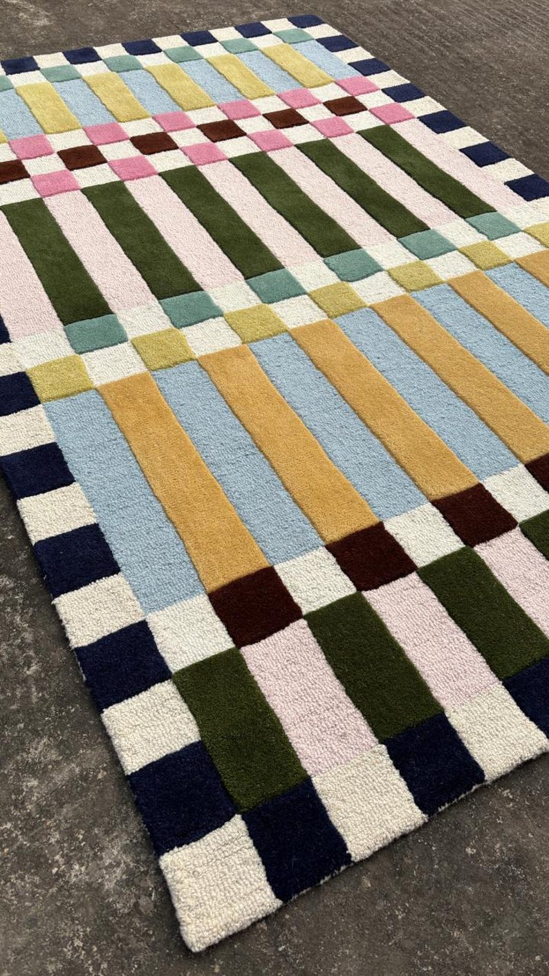 May include: A rectangular area rug featuring a geometric pattern of alternating vertical stripes and squares. The design incorporates a palette of navy blue, olive green, mustard yellow, light blue, pale pink, cream, and lime green tufted textures.