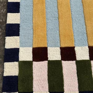 May include: A tufted rug featuring a geometric pattern of rectangular blocks in navy blue, cream, olive green, light pink, mustard yellow, light blue, and dark brown. The design consists of a vertical striped center bordered by a checkered edge.