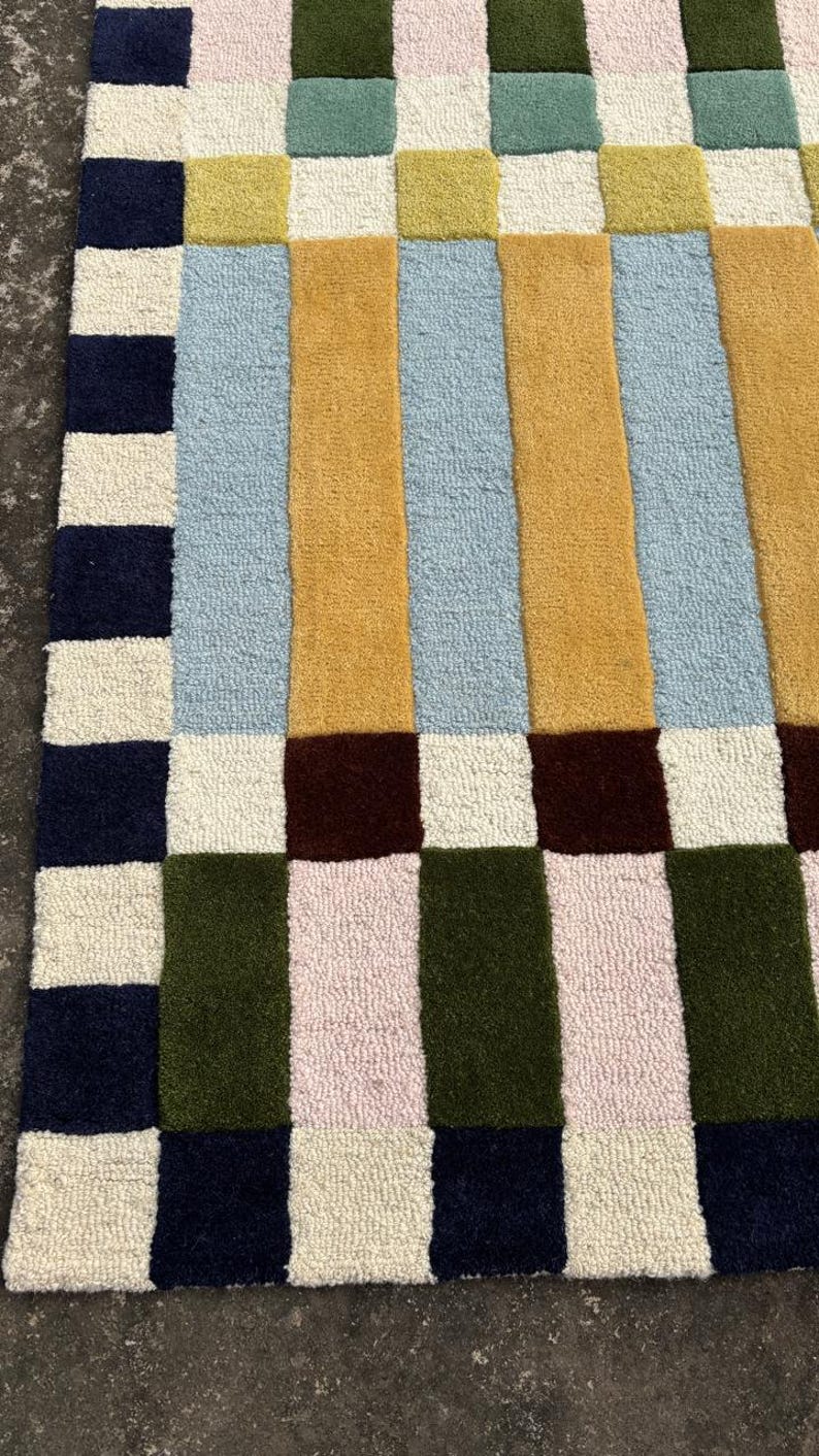 May include: A tufted rug featuring a geometric pattern of rectangular blocks in navy blue, cream, olive green, light pink, mustard yellow, light blue, and dark brown. The design consists of a vertical striped center bordered by a checkered edge.