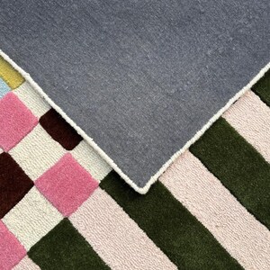 May include: A tufted rug featuring a geometric pattern of pink, dark brown, and light pink squares alongside alternating dark green and light pink stripes. The rug is folded over to reveal a solid dark gray fabric backing with white stitched edges.