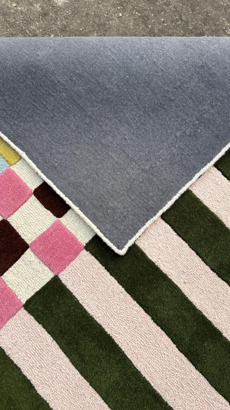 May include: A tufted rug featuring a geometric pattern of pink, dark brown, and light pink squares alongside alternating dark green and light pink stripes. The rug is folded over to reveal a solid dark gray fabric backing with white stitched edges.