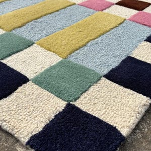 May include: A tufted area rug featuring a geometric grid pattern composed of rectangular and square blocks in various colors including navy blue, cream, light blue, olive green, mustard yellow, pink, and dark brown with a textured pile surface.