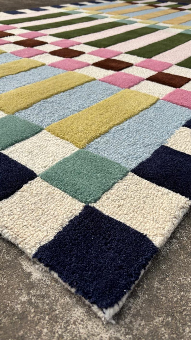 May include: A tufted area rug featuring a geometric grid pattern composed of rectangular and square blocks in various colors including navy blue, cream, light blue, olive green, mustard yellow, pink, and dark brown with a textured pile surface.