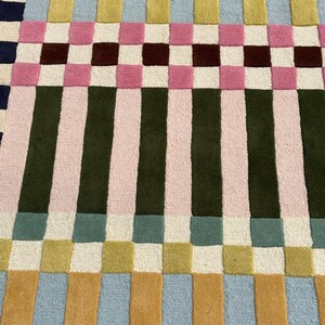 May include: A rectangular area rug featuring a geometric pattern of alternating vertical stripes and squares. The design incorporates a palette of navy blue, light blue, mustard yellow, olive green, soft pink, maroon, and cream in a grid layout.