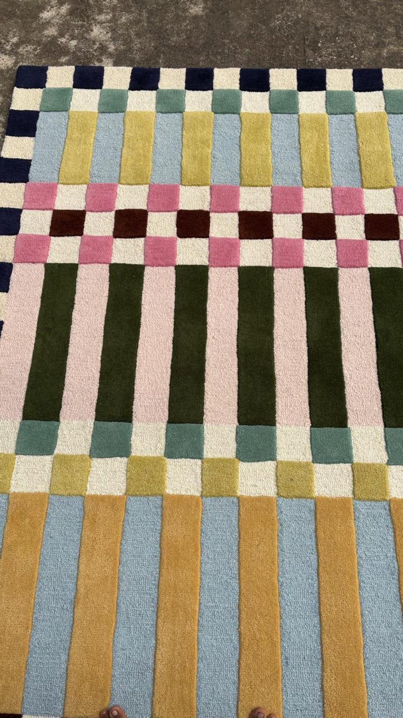 May include: A rectangular area rug featuring a geometric pattern of alternating vertical stripes and squares. The design incorporates a palette of navy blue, light blue, mustard yellow, olive green, soft pink, maroon, and cream in a grid layout.