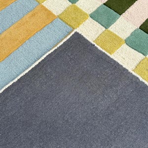 May include: A tufted rug featuring a geometric pattern of squares and stripes in shades of dark blue, yellow, light blue, cream, sage green, and dark olive green. The underside of the rug is visible, showing a solid dark gray fabric backing.