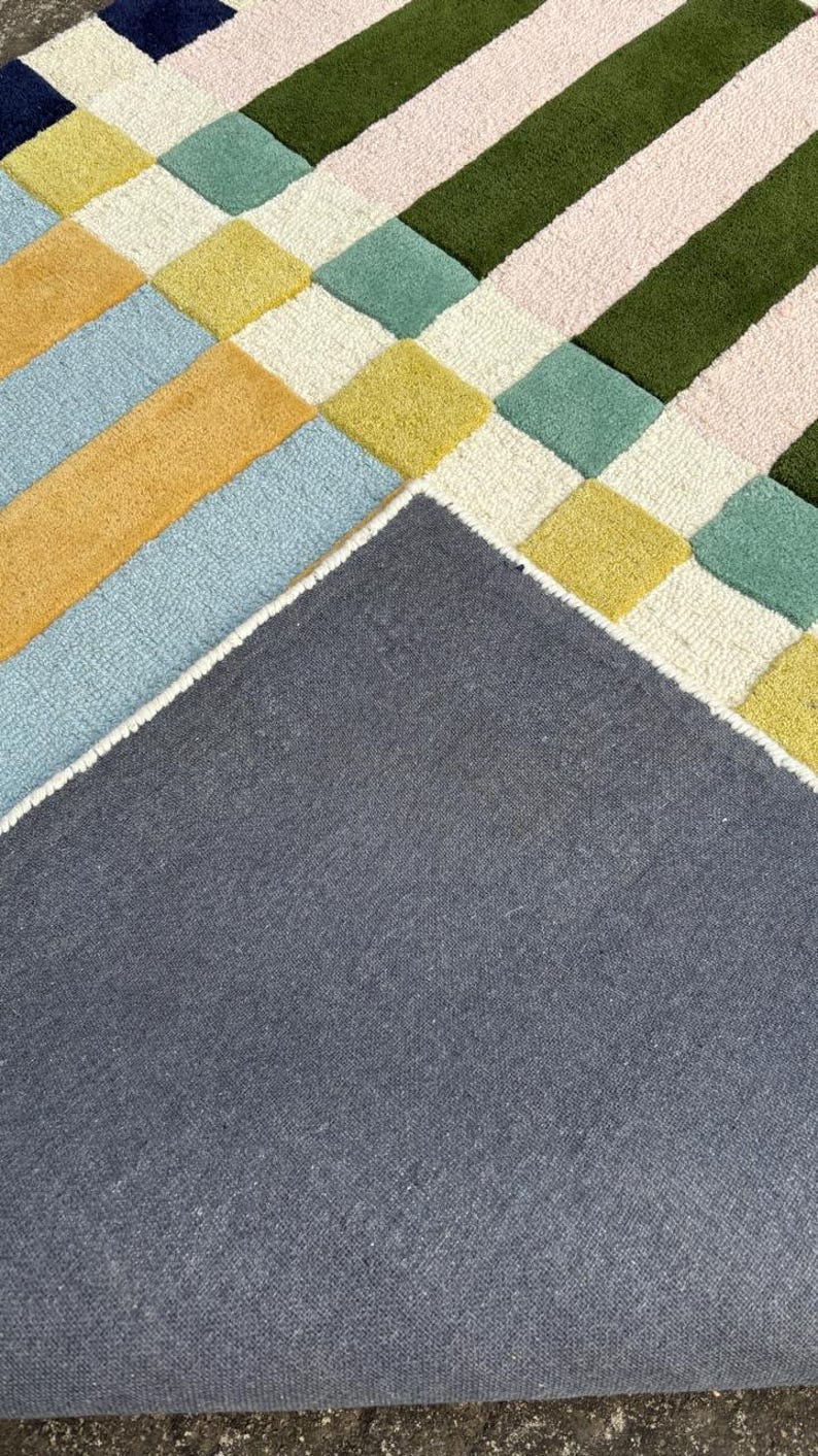 May include: A tufted rug featuring a geometric pattern of squares and stripes in shades of dark blue, yellow, light blue, cream, sage green, and dark olive green. The underside of the rug is visible, showing a solid dark gray fabric backing.