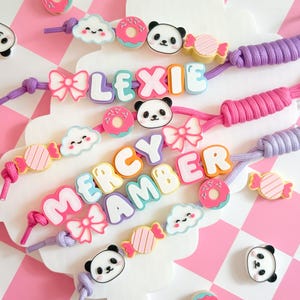 May include: Three custom lanyards featuring colourful silicone beads shaped like pandas, donuts, clouds, bows, and candies. The lanyards display the names LEXIE, MERCY, and AMBER in chunky, multi-coloured block letters, strung on purple and pink cords.