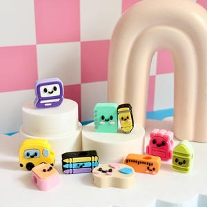 May include: A collection of small, colorful, kawaii-style resin charms featuring school-themed designs including a purple computer, a mint green notebook, a yellow pencil, a yellow school bus, a pink backpack, crayons, and a paint palette.