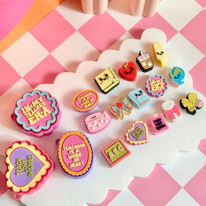 May include: A collection of colorful, small, decorative charms featuring school-themed designs, including hearts, books, pencils, and globes. Several pieces display text such as "In My Teacher Era" and "Teaching Is A Work Of Heart" in various fonts.