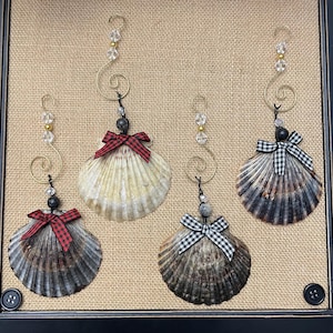 May include: Four hanging seashell ornaments featuring gold-tone metal spirals, clear and gold beads, and small fabric bows. Two shells are dark grey with red and black plaid bows, one is light cream with a red plaid bow, and one is dark with a black and white gingham bow.
