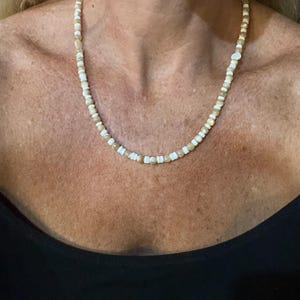 May include: A beaded necklace featuring a repeating pattern of small, round, translucent beige beads and flat, white, disc-shaped beads. The necklace is strung on a thin cord and rests against the neck, showcasing a simple, uniform design.