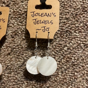 May include: A pair of dangle earrings featuring circular white mother-of-pearl discs suspended from silver-toned metal hooks. The earrings are attached to a tan cardboard display card with the handwritten text JOLEAN'S JEWELS ~JJ printed on it.