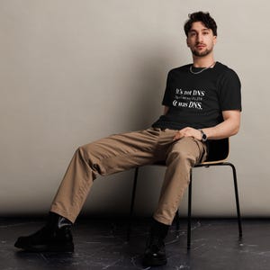 Op de afbeelding: Een zwart T-shirt met korte mouwen en witte tekst: It's not DNS. There's no way it's DNS. It was DNS. Het T-shirt wordt gecombineerd met een beige broek en zwarte laarzen, wat zorgt voor een casual uitstraling met een humoristische technische print.