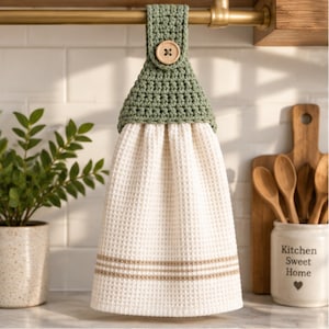 May include: A hanging kitchen towel featuring a cream-coloured waffle-weave fabric with horizontal tan stripes. The top section is made of sage green crocheted yarn and is secured with a single round wooden button to a gold-toned metal rod.