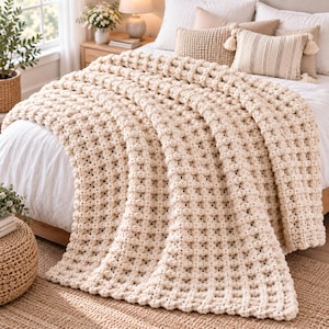 May include: A cream-colored chunky knit throw blanket with a raised waffle-like texture. The blanket features a thick, soft yarn construction with a repeating square grid pattern, draped over the foot of a bed with matching pillows.