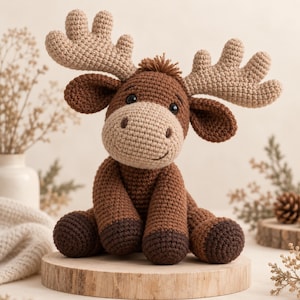 May include: A crocheted moose stuffed animal featuring a dark brown body, a lighter tan muzzle, and large beige antlers. The toy has small dark brown hooves, black safety eyes, and a small tuft of brown hair on top of its head, sitting in a seated position.