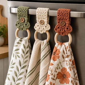 May include: Three hanging kitchen towels with crocheted tops in olive green, cream, and terracotta. Each towel features a wooden ring, a matching crocheted tab with a brown button, and distinct patterns: green leaves, green stripes, and orange flowers.