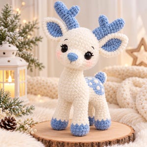 Puede incluir: Un peluche de ciervo tejido a ganchillo con cuerpo color crema y astas, orejas, pezuñas y cola moteada en azul claro. La figura tiene grandes ojos negros, una pequeña nariz azul y mejillas rosadas, de pie sobre sus cuatro patas.