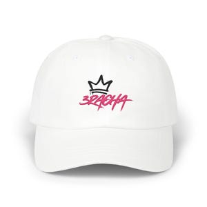 May include: A white baseball cap featuring a black embroidered crown icon positioned above the pink embroidered text 3RACHA. The cap has a structured front panel, a curved brim, and a button at the top center of the crown, presenting a clean design.