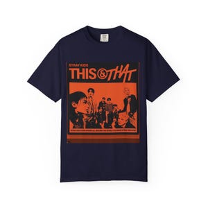 May include: A navy blue short-sleeve t-shirt featuring a large rectangular graphic in orange and black. The graphic displays the text "STRAY KIDS THIS & THAT" and "STRAY KIDS EVERYWHERE ALL AROUND THE WORLD YOU MAKE STRAY KIDS STAY" with a group of people.