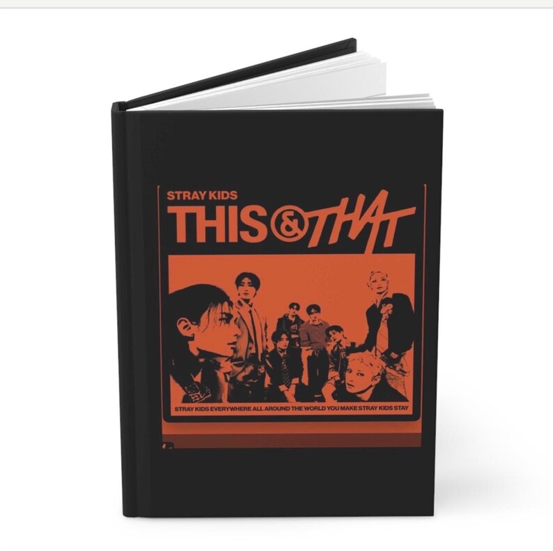 May include: A black hardcover book featuring an orange graphic on the front cover. The text reads STRAY KIDS THIS & THAT and STRAY KIDS EVERYWHERE ALL AROUND THE WORLD YOU MAKE STRAY KIDS STAY. The graphic includes a stylized illustration of a group.