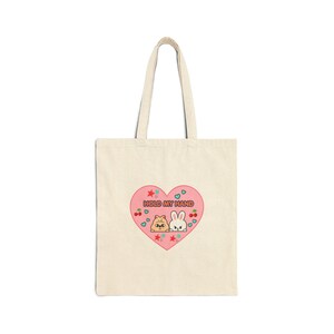 May include: A beige canvas tote bag featuring a pink heart graphic in the center. Inside the heart, the text "HOLD MY HAND" appears above illustrations of a small brown creature and a white rabbit, surrounded by tiny red stars, cherries, and blue hearts.