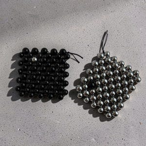 May include: Two square-shaped beaded mats constructed from interconnected spherical beads. One mat features glossy black beads with a single silver bead, while the other consists of metallic silver beads. Both mats are strung together with thin black cord.