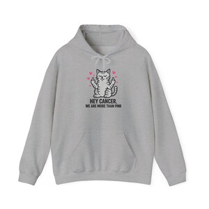 May include: A heather gray hooded sweatshirt featuring a graphic of a gray tabby cat with its eyes closed, surrounded by small pink hearts, and giving the middle finger with both paws. Below the cat is the text: HEY CANCER, WE ARE MORE THAN PINK.