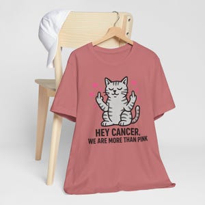 May include: A dusty rose t-shirt featuring a graphic of a grey tabby cat with closed eyes, holding up its middle fingers on both hands. Small pink hearts surround the cat. The text below reads, HEY CANCER, WE ARE MORE THAN PINK.