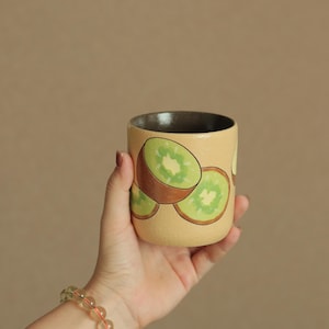 May include: A cylindrical ceramic cup with a textured beige exterior featuring a hand-painted pattern of sliced green kiwi fruit. The interior of the cup is a smooth, dark brown finish. The vessel has a straight rim and a flat base.