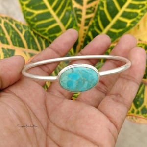 May include: A silver cuff bracelet featuring a smooth, polished metal band and a central oval-shaped turquoise gemstone with natural veining. The stone is set in a simple silver bezel, creating a clean, balanced design for the wrist.
