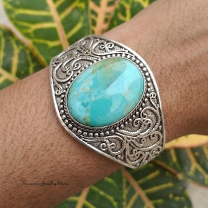 May include: A silver cuff bracelet featuring a large, oval-shaped turquoise stone with natural veining. The metal band is decorated with intricate, raised scrollwork patterns and a beaded border surrounding the central gemstone setting.