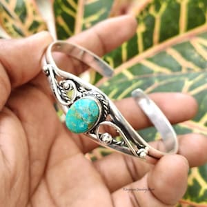 May include: A silver cuff bracelet featuring a central oval turquoise stone with brown matrix veining. The metal band displays decorative twisted wire detailing and smooth, curved silver accents. The text Kingman Jewelry Store is visible.