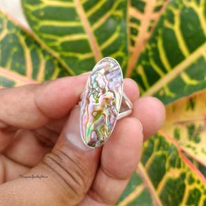May include: A silver ring featuring a large, oval-shaped abalone shell inlay. The shell displays a vibrant, iridescent surface with swirling patterns of pink, green, blue, and purple hues. The metal band is smooth and polished, holding the stone.