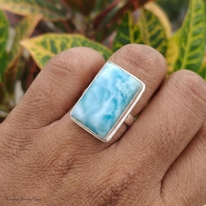 May include: A rectangular Larimar gemstone set in a polished silver ring. The stone features a marbled pattern of sky blue and white tones with a smooth, flat surface. The silver band is simple and holds the stone securely in a bezel setting.